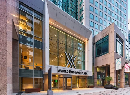 A sleek, modern building with a glass facade reflecting light. The entrance is illuminated, and there are people walking on the sidewalk in front. The setting conveys a bustling urban environment.