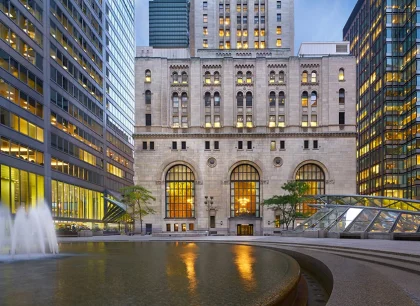 A historic building with ornate architecture contrasted by modern skyscrapers on either side. The foreground features a large, round water fountain. The area is well-lit, creating a lively urban atmosphere.