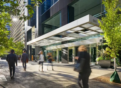 The entrance of a modern office building with a glass canopy. People are walking on the sidewalk, and there are trees and greenery around, creating a lively urban atmosphere.