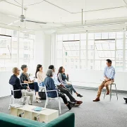 A group of people seated in a bright, modern room, engaged in a meeting or presentation. One person is speaking while the others listen.