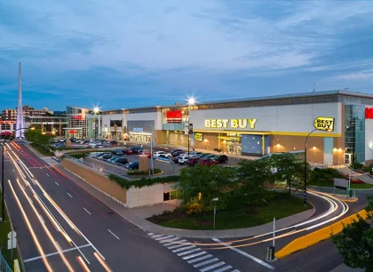Un grand centre commercial avec plusieurs magasins et une aire de stationnement achalandée, représenté au crépuscule par des enseignes illuminées.