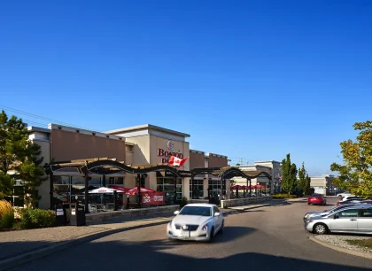 A shopping plaza with parked cars in front of stores under a clear blue sky. The area is bustling and well-maintained.