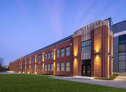 A modern brick building with large windows and exterior lighting, set against a twilight sky.