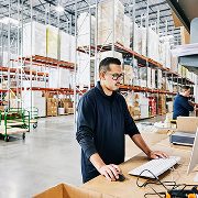 Personne travaillant sur un ordinateur portable dans un entrepôt spacieux rempli de tablettes et de boîtes.