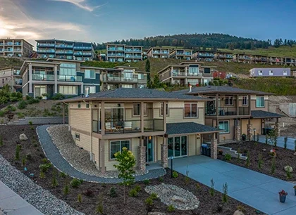 A modern, stylish house with large windows and landscaped surroundings, situated in a residential area with hills in the background at dusk.